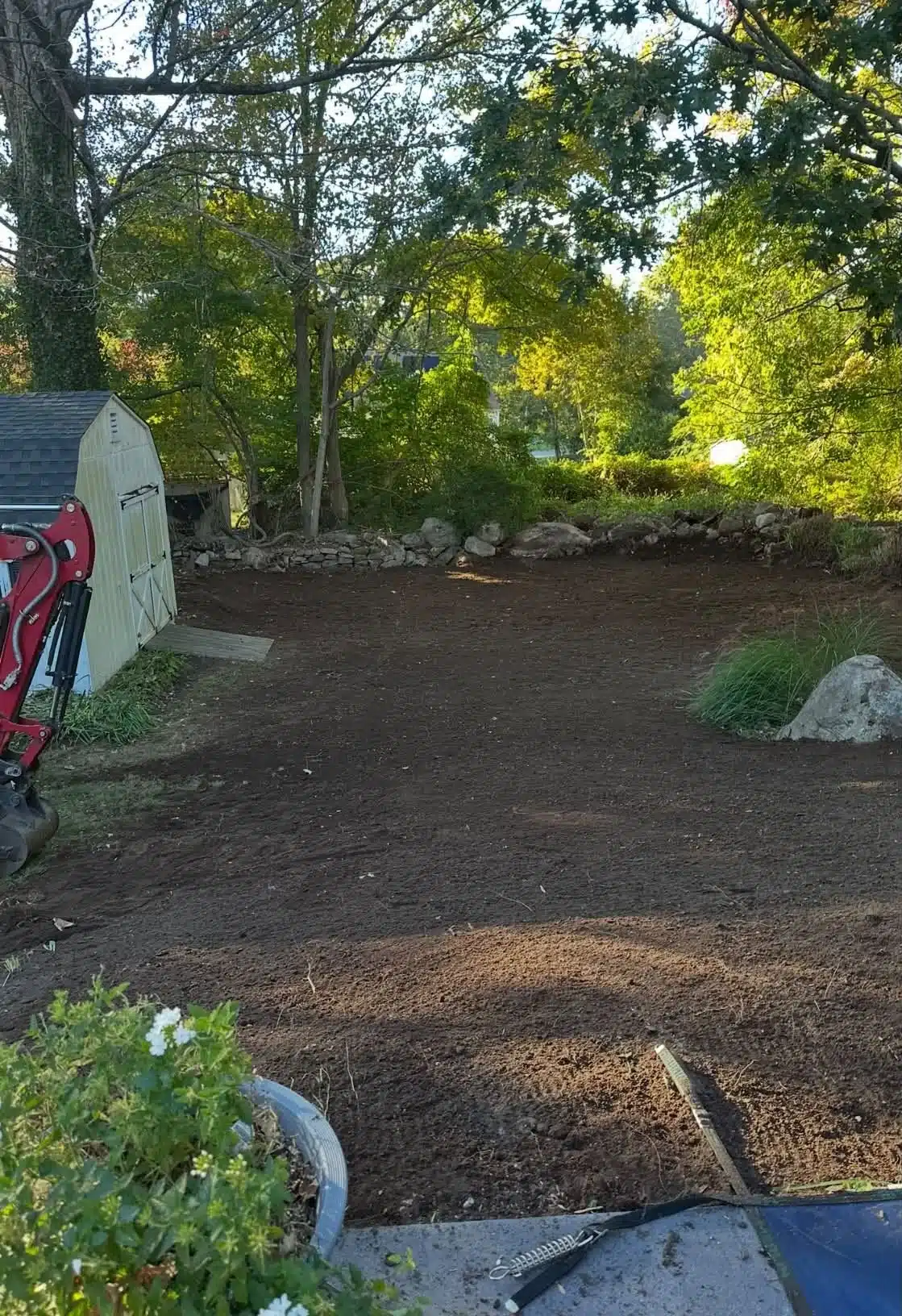 Residential brush clearing in Ledyard, CT, showing a cleared yard with freshly prepared soil, natural stone borders, and a storage shed surrounded by trees.