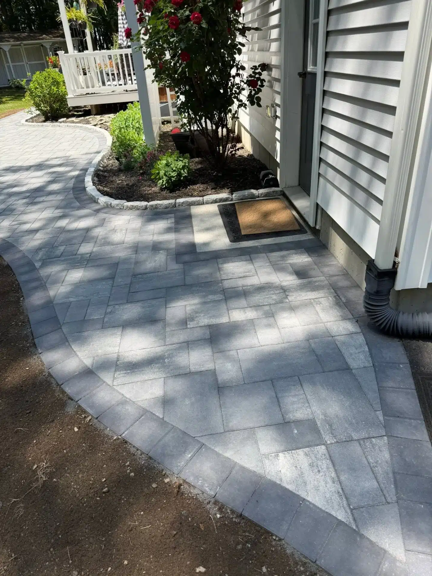 hardscaping-services-waterford-ct-spicers-lawn-care (1) Custom stone paver patio hardscaping in Waterford, CT, with a curved layout and decorative garden beds beside the home.